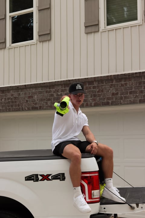 Man sitting on white pickup truck bed wearing white shirt, black shorts, and cap, holding yellow binoculars in front of garage