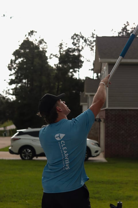 Man in blue shirt cleaning gutters on brick house with extended pole, trees and driveway visible in background