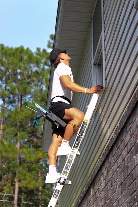 Man climbing a white ladder against a metal building wall on a sunny day with trees in the background